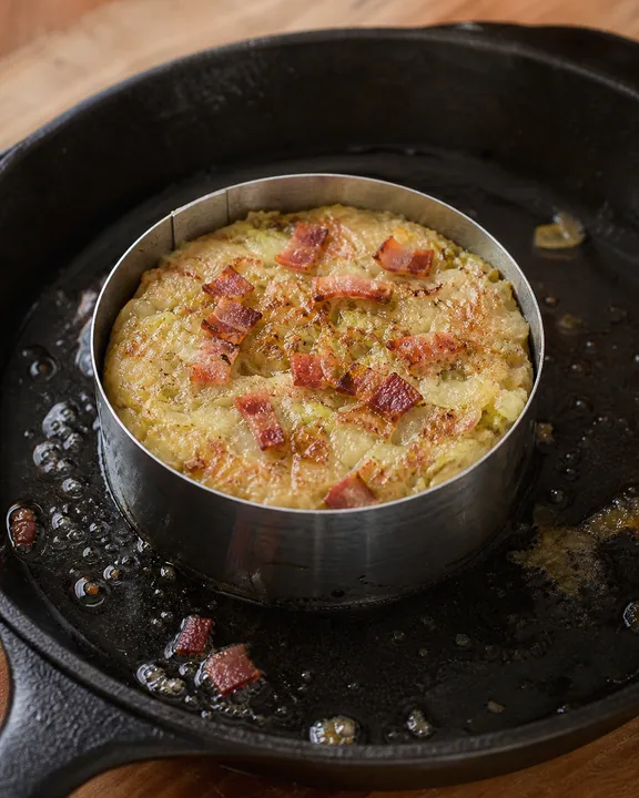 Porción individual de trinxat friéndose en un aro con bacon