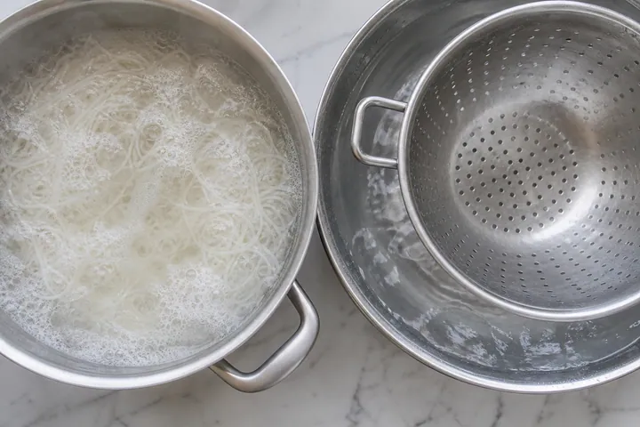 Thin rice noodles cooking in a pot beside a colander