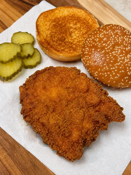 Pollo frito escurriendo junto a panecillo y rodajas de pepinillo
