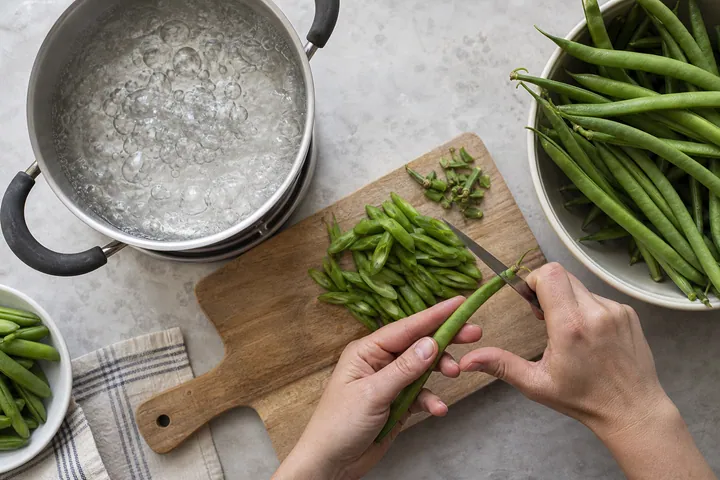 Hands trimming green runner beans on a cutting board beside boiling water