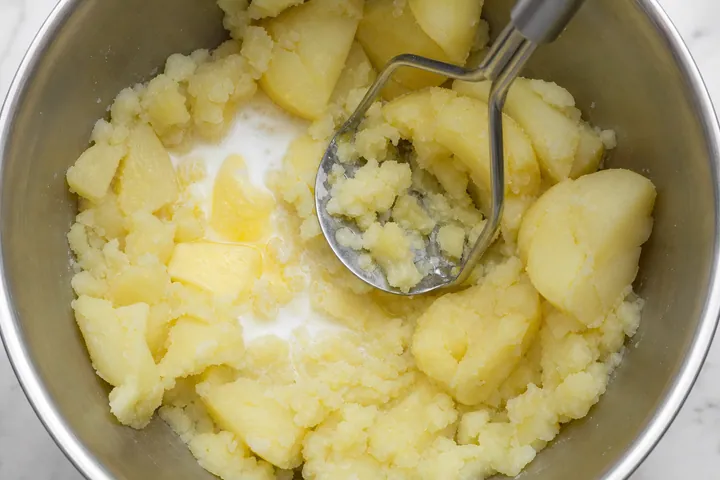 A potato masher mashing boiled potatoes with butter and milk in a bowl